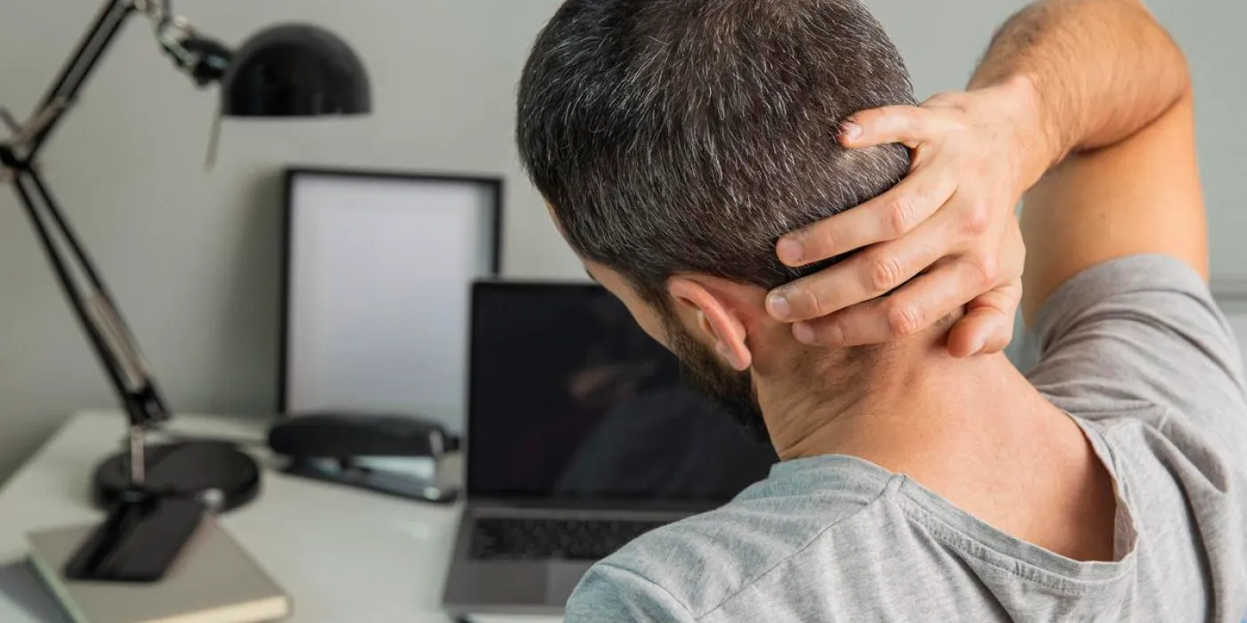 Homem sentado em frente ao computador sentindo dor no pescoço.