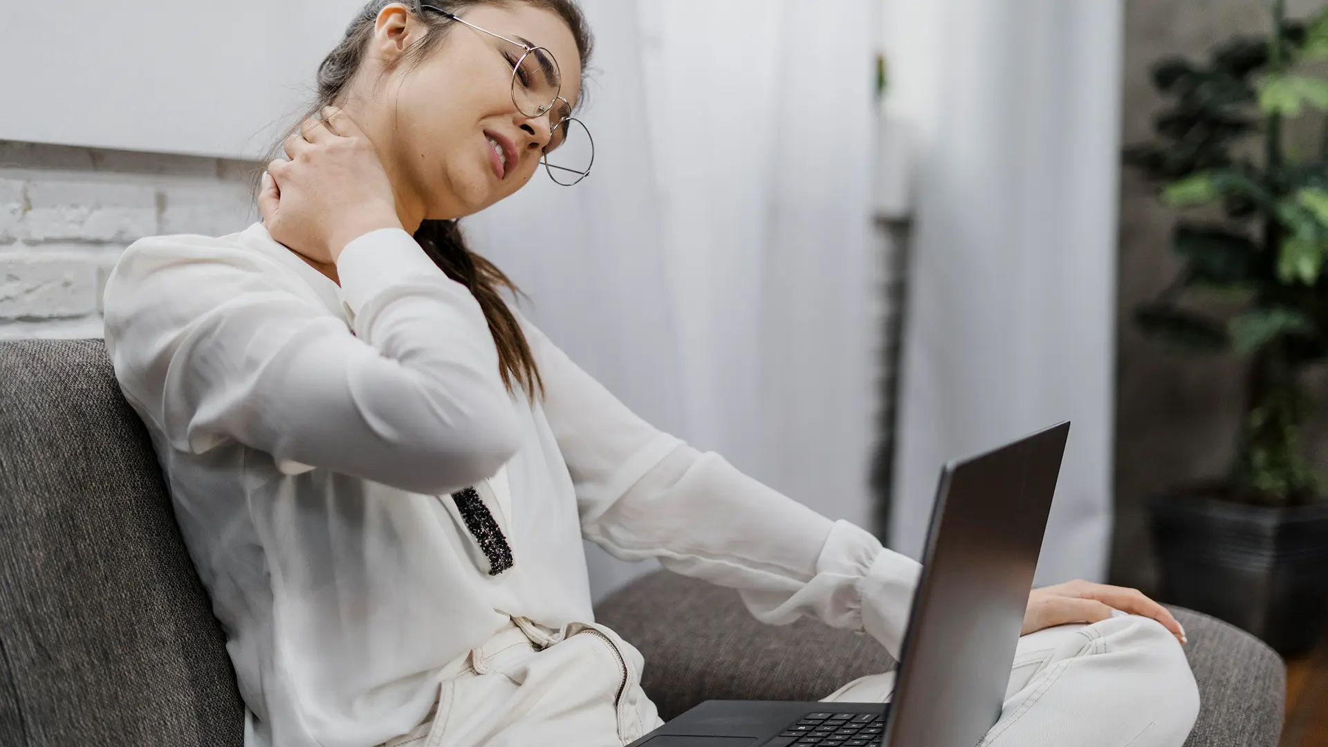 Mulher sentada com notebook no colo enquanto põe a mão na nuca, indicando sentir dor